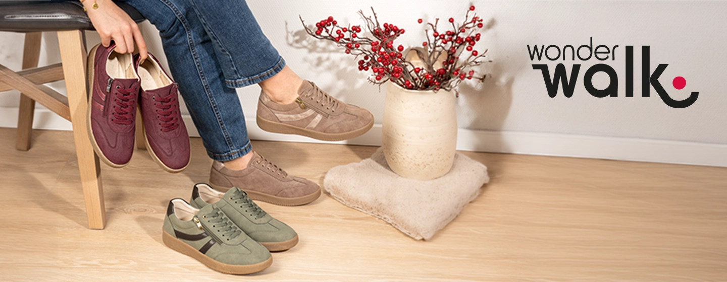 Eine Person, die auf einem Stuhl sitzt, hält burgunderrote Schuhe in der Hand, trägt beigefarbene Schuhe, während grüne Schuhe auf dem Boden liegen, neben roten Beeren in einer Vase und dem „Wonder Walk“-Logo.