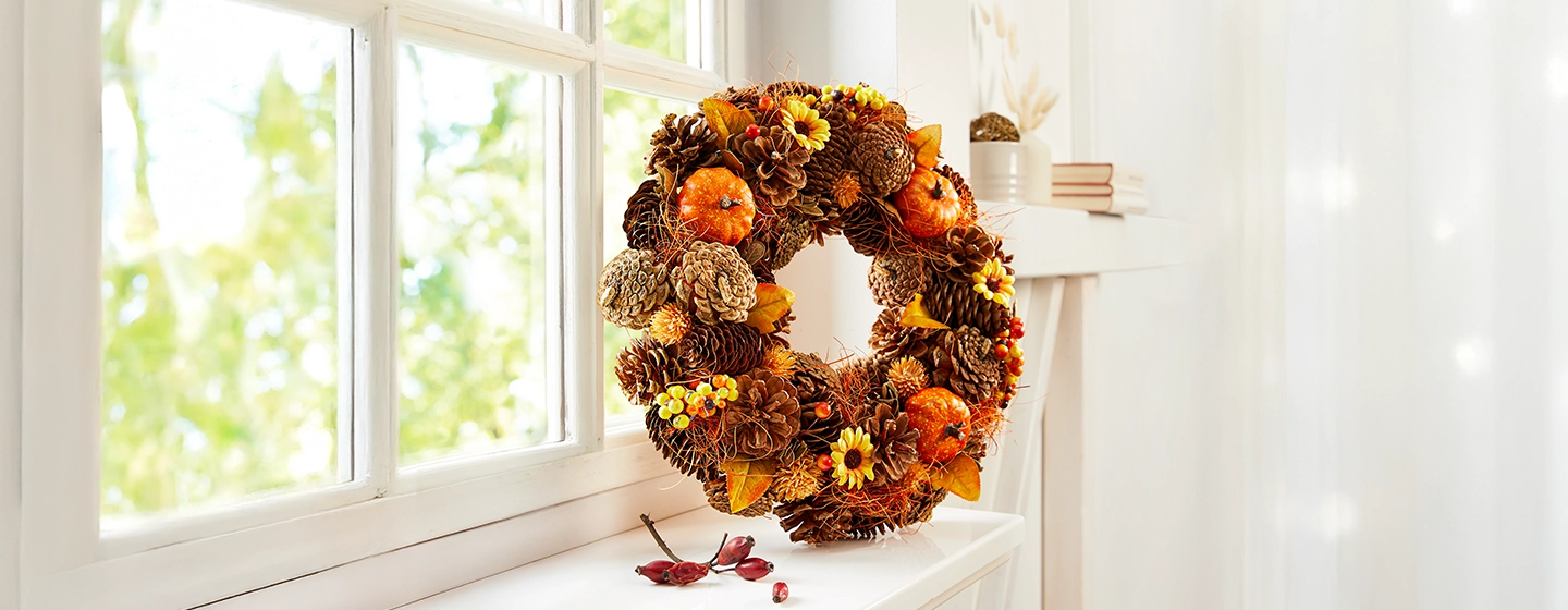 Auf einer weißen Fensterbank liegt ein Herbstkranz mit Tannenzapfen, Minikürbissen, Beeren und Sonnenblumen, während Licht durch das Fenster hereinströmt.