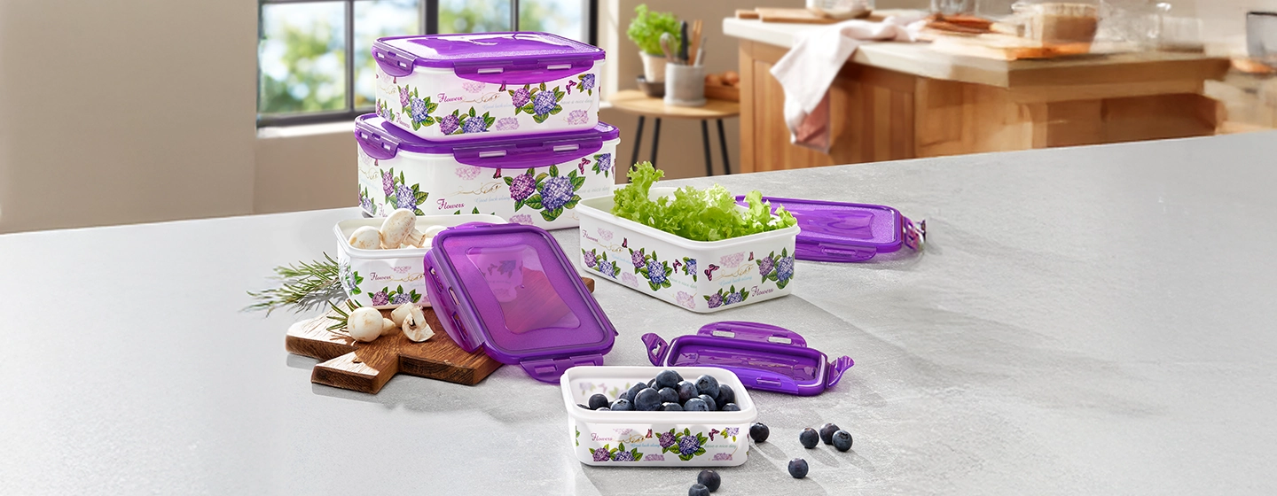 Küchenzeile mit Behältern mit violetten Deckeln und Blumenmuster, in denen Blaubeeren, Salat und Knoblauch aufbewahrt werden. Im Hintergrund eine sonnendurchflutete Küche.