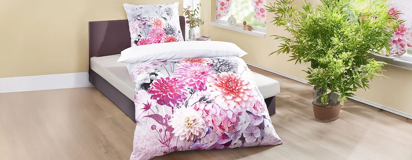 Ein ordentlich gemachtes Bett mit Bettwäsche mit Blumenmuster in einem modernen Zimmer mit hellem Holzboden, einer Topfpflanze und großen Fenstern.