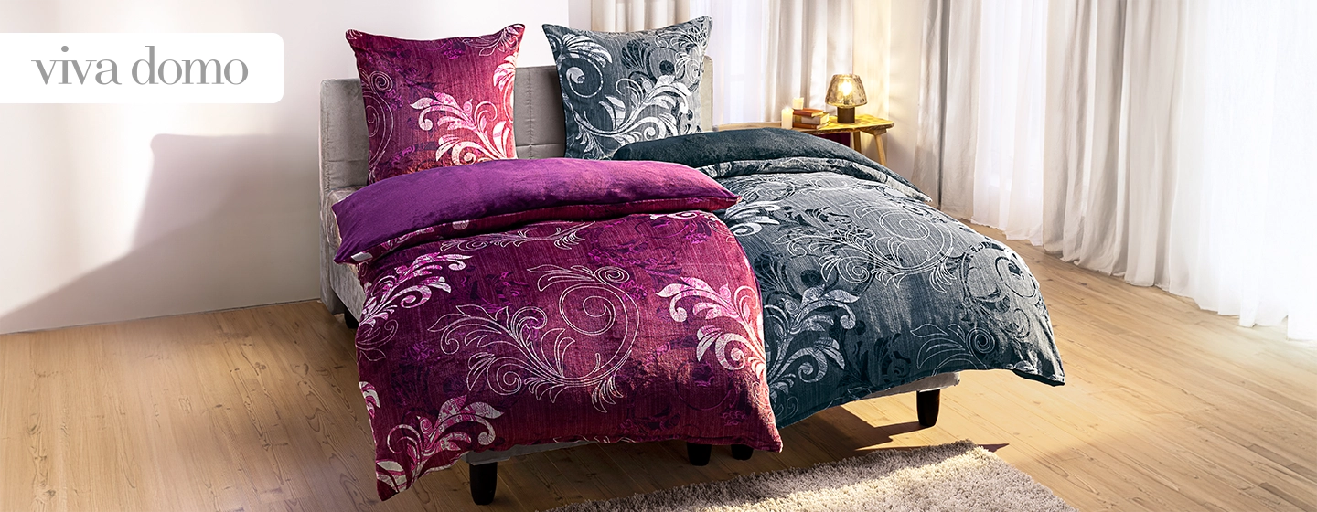 Ein Schlafzimmer mit einem Bett, das mit lebhaften lila-grauen Bettwäsche mit Blumenmuster bezogen ist, vor transparenten Vorhängen und einem Holzboden.
