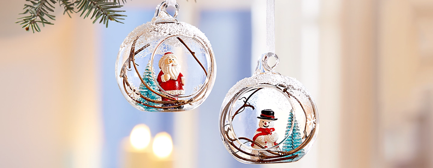 An einem Baum hängen zwei gläserne Weihnachtskugeln, eine mit einer Weihnachtsmannfigur und die andere mit einem Schneemann, beide umgeben von verschneiten Zweigen.