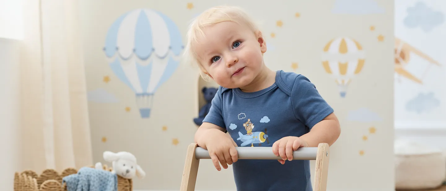 Ein Baby mit blonden Haaren lehnt sich an einen hölzernen Lauflernwagen, trägt einen blauen Strampler; im Hintergrund sind Wanddekorationen mit Heißluftballons zu sehen.