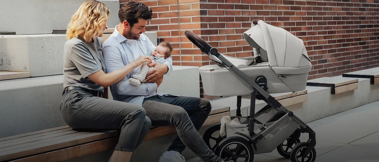 Ein Paar sitzt auf einer Bank und lächelt sein Baby an. Der Vater hält das Baby im Arm, daneben steht ein Kinderwagen. Es ist ein sonniger Tag.