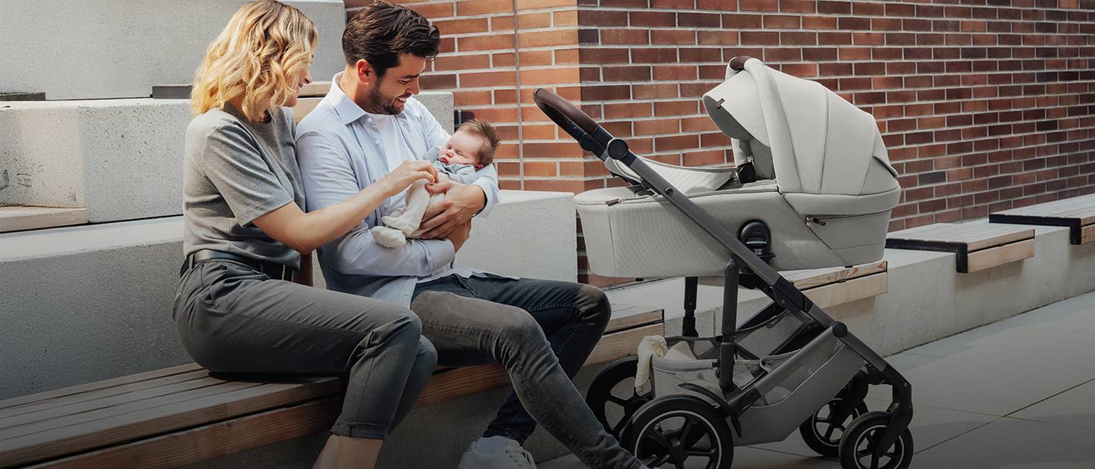 Ein Paar sitzt auf einer Bank und lächelt sein Baby an. Der Vater hält das Baby im Arm, daneben steht ein Kinderwagen. Es ist ein sonniger Tag.