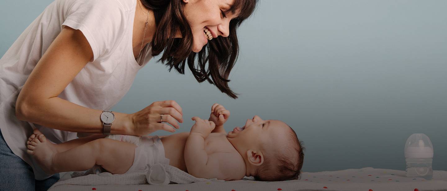 Eine Frau lächelt ein lachendes Baby an, das auf einem Wickeltisch liegt und in dessen Nähe eine Babyflasche steht.