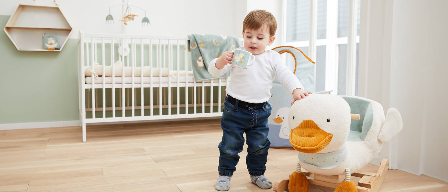 Ein Kleinkind mit einer Tasse steht neben einem Schaukelspielzeug in Entenform in einem Kinderzimmer mit Kinderbett und weicher Einrichtung.