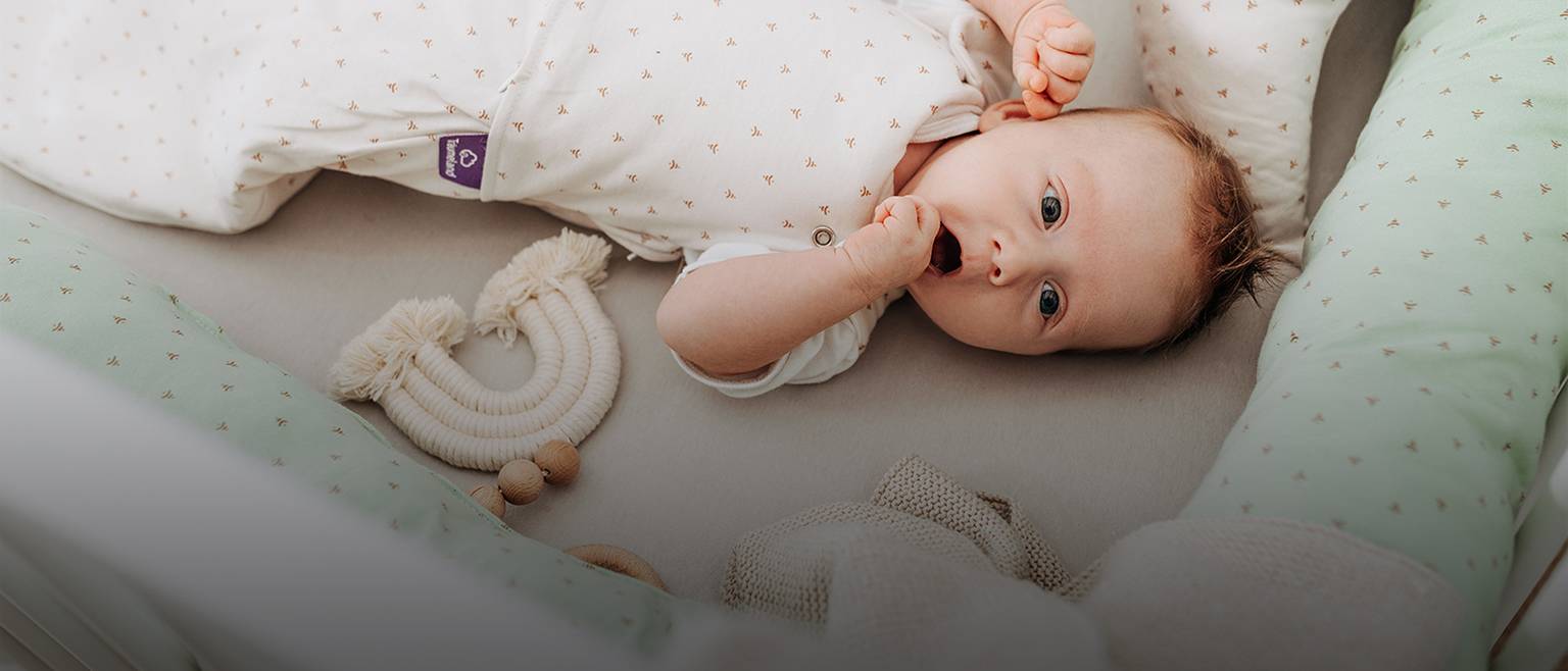 Baby liegt in einem Kinderbett, trägt einen weißen Strampler mit kleinen Mustern und ist von weicher mintgrüner Bettwäsche und Spielzeug umgeben.