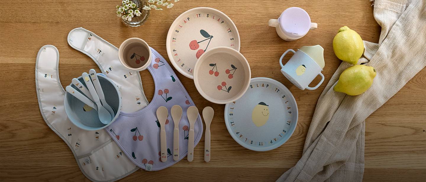 Buntes Kinder-Essservice mit Tellern, Schüsseln, Tassen und Besteck mit Fruchtmotiven, daneben Zitronen und eine Tischdecke auf einem Holztisch.