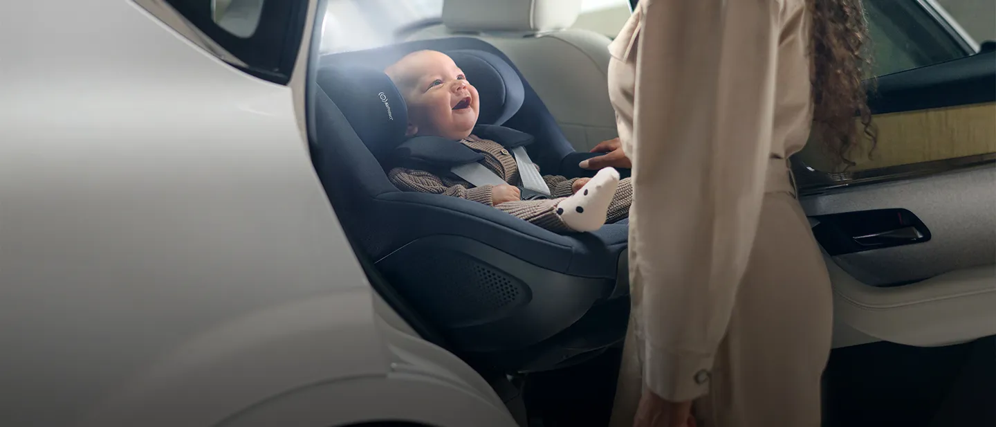 Ein lächelndes Baby in einem Autositz blickt zu einem Erwachsenen in einem Auto auf.