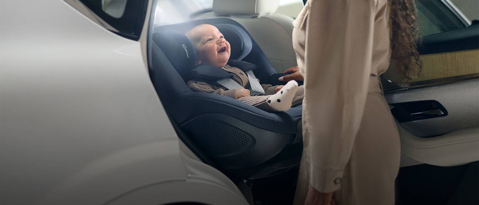Ein lächelndes Baby in einem Autositz blickt zu einem Erwachsenen in einem Auto auf.