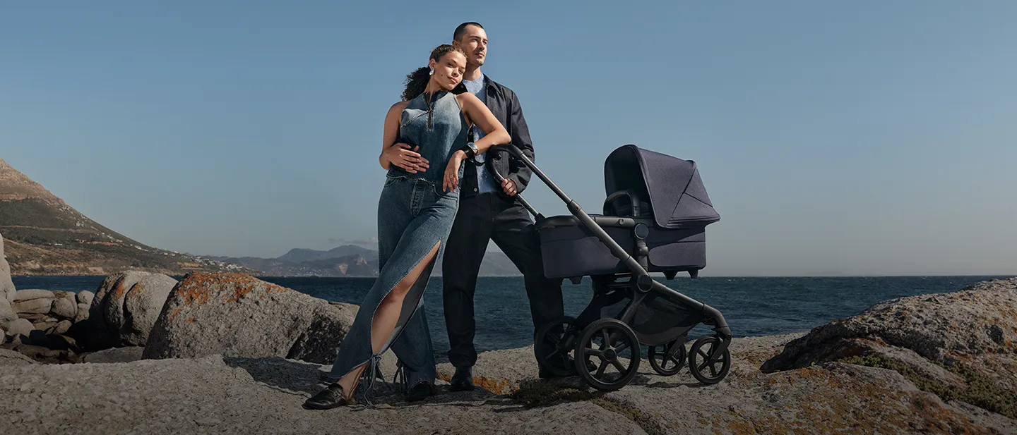 Paar in Jeanskleidung steht am felsigen Ufer mit einem marineblauen Kinderwagen, Meer und Berge im Hintergrund unter einem klaren blauen Himmel.