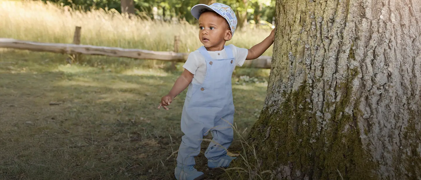 Ein Kleinkind mit blauer Mütze und Latzhose steht in einem sonnenbeschienenen Park an einem Baum, eine Hand am Stamm, im Hintergrund Gras.