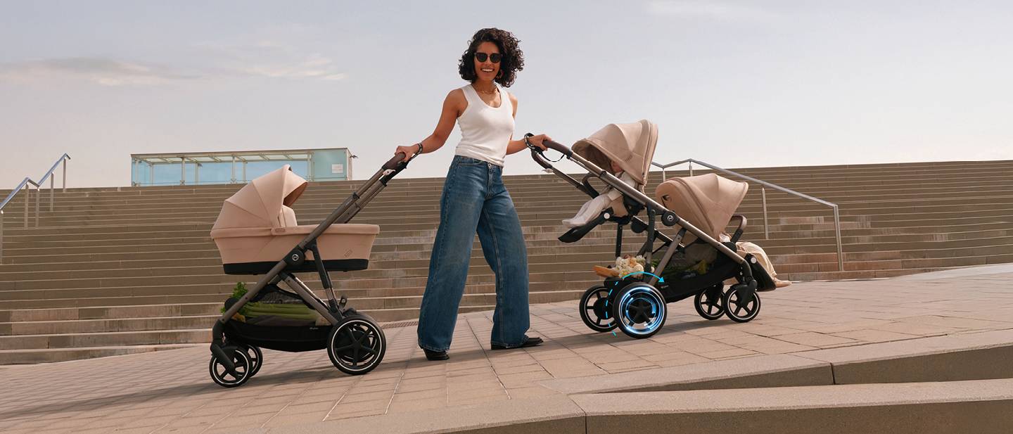 Eine Frau in einem weißen Tanktop und Jeans schiebt zwei beige Kinderwagen auf einem gepflasterten Platz; im Hintergrund sind Treppen zu sehen.
