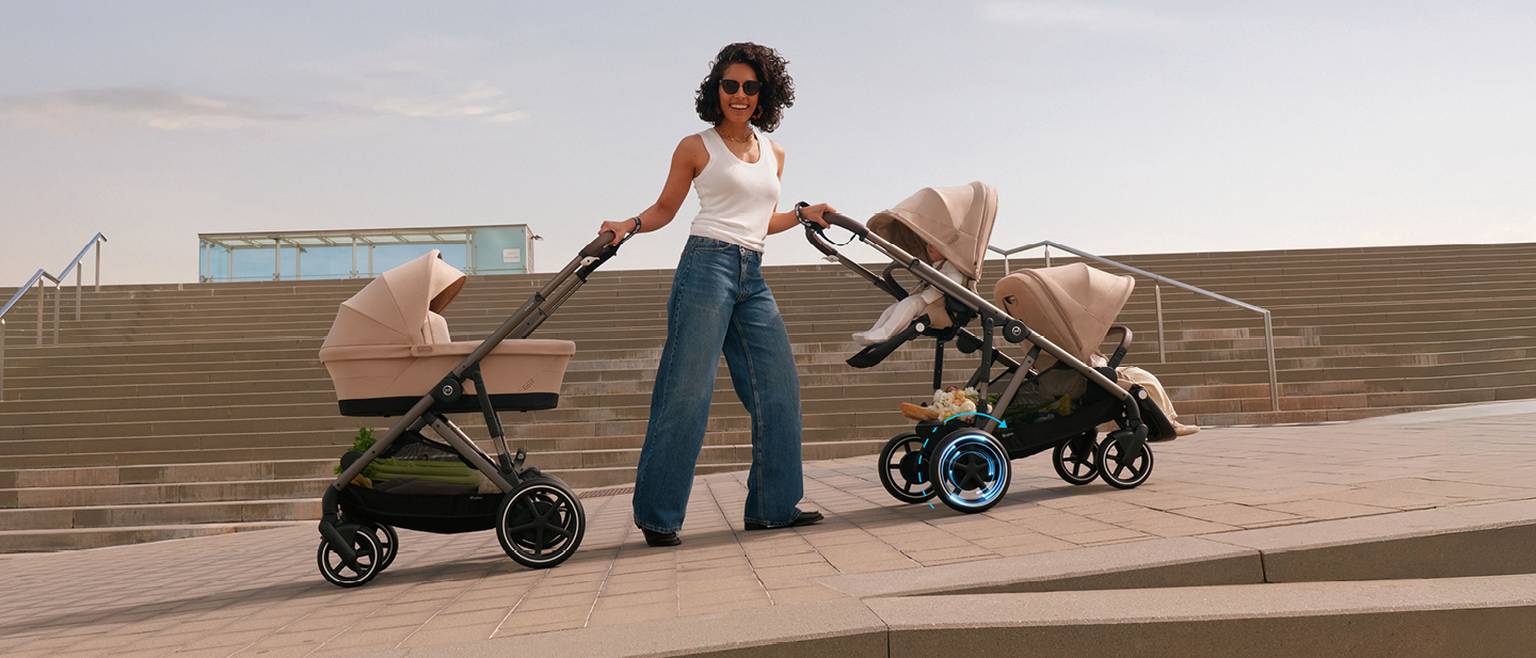 Eine Frau in einem weißen Tanktop und Jeans schiebt zwei beige Kinderwagen auf einem gepflasterten Platz; im Hintergrund sind Treppen zu sehen.