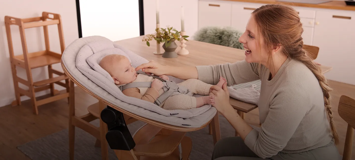 Eine Frau lächelt und interagiert mit einem Baby, das in einem gepolsterten Sitz auf einem Esstisch in einem gemütlichen Zimmer liegt.