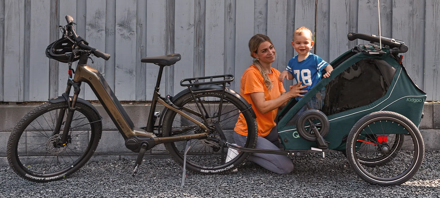 Eine Frau kniet neben einem Fahrrad mit Anhänger, in dem sich ein lächelndes Kind befindet; beides steht in der Nähe einer Holzmauer auf Kies.