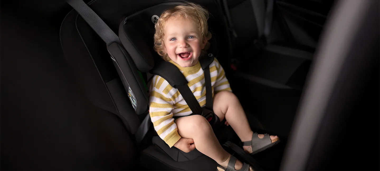Ein lächelndes Kleinkind mit lockigem Haar sitzt in einem Kindersitz auf dem Rücksitz eines Fahrzeugs. Es trägt einen gestreiften Pullover und Sandalen.