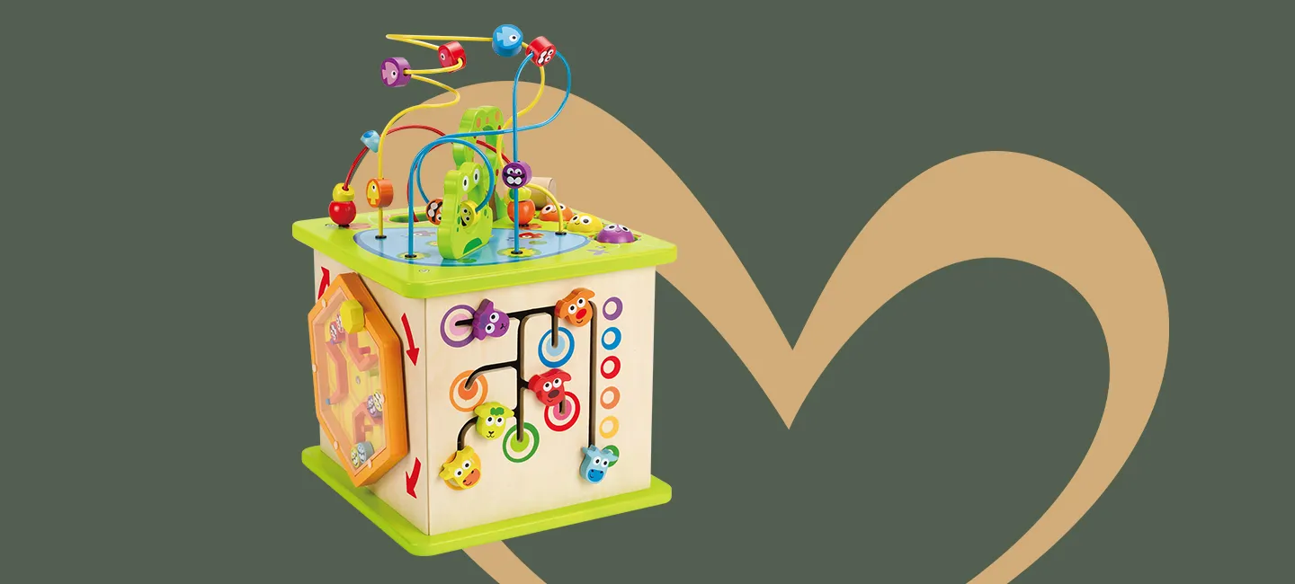 Buntes Aktivitätswürfel-Spielzeug aus Holz mit Perlenlabyrinth, Zahnrädern, Formensortierern und Knöpfen vor grünem Hintergrund mit einem hellbraunen Herz.