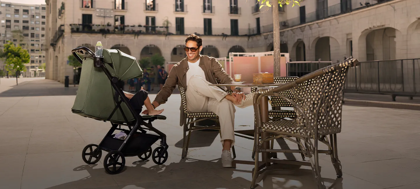 Ein Mann mit Sonnenbrille sitzt in einem Straßencafé und schiebt einen Kinderwagen mit einem Baby unter einem grünen Baldachin vor dem Hintergrund einer Stadtkulisse.