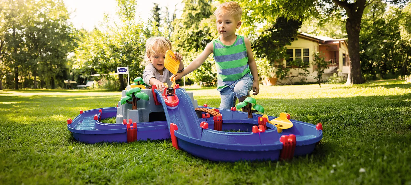 Zwei Kinder spielen auf einer sonnigen Rasenfläche mit einer bunten Wasserbahn, umgeben von Bäumen und einem Haus im Hintergrund.