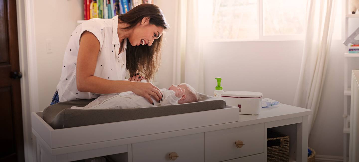 Eine Frau lächelt ein Baby auf einem Wickeltisch in einem hellen Raum mit Regalen und einem Fenster an.