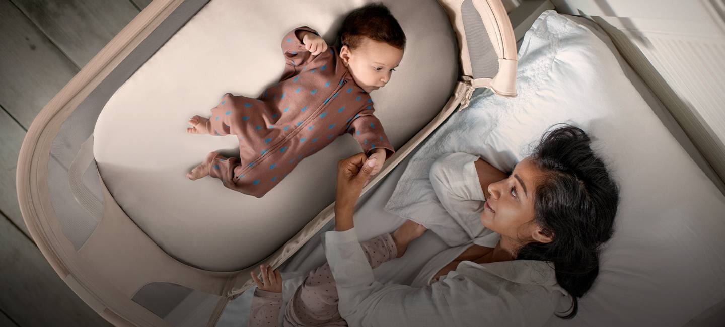 Eine Mutter liegt im Bett und hält ein Baby in der Wiege an der Hand. Das Baby trägt einen braunen Strampler mit blauen Punkten. Tageslicht durchflutet den Raum.