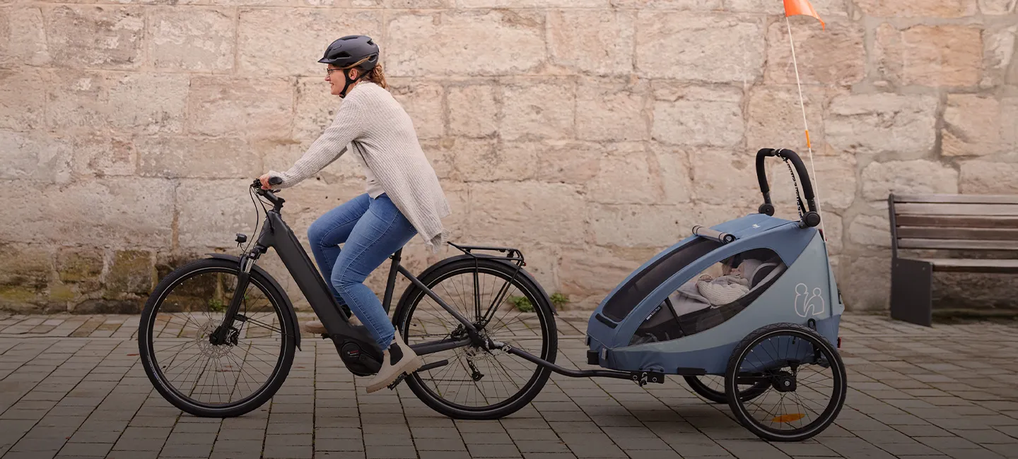 Eine Frau fährt mit dem Fahrrad und zieht ein Kind in einem blauen Anhänger auf einem gepflasterten Weg neben einer Steinmauer.