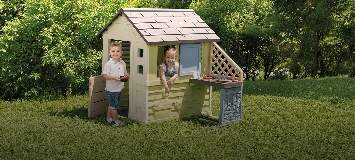 Zwei Kinder spielen in einem Garten mit einem Spielhaus. Eines steht draußen, das andere sitzt im Fenster, umgeben von Grün.
