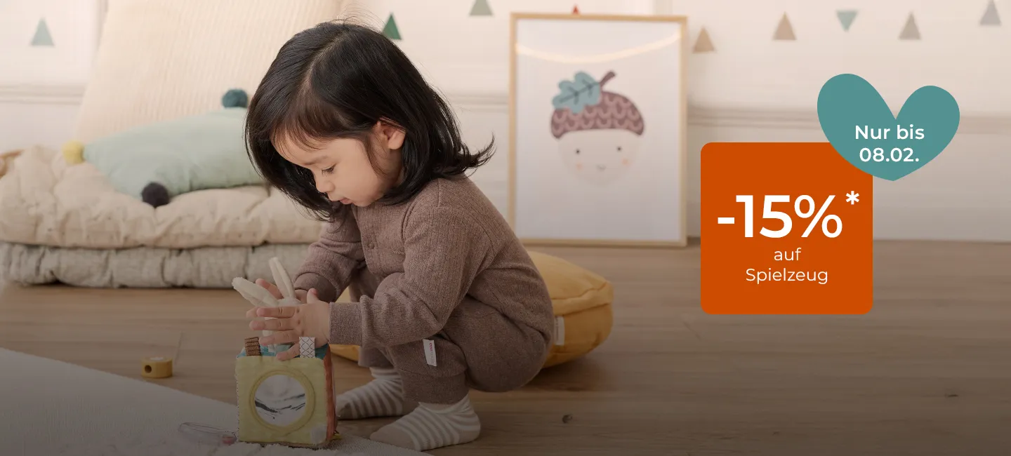Kleinkind spielt in einem gemütlichen Zimmer mit einem Bauklotz. (Text bietet bis zum 8. Februar 15 % Rabatt auf Spielzeug.)
