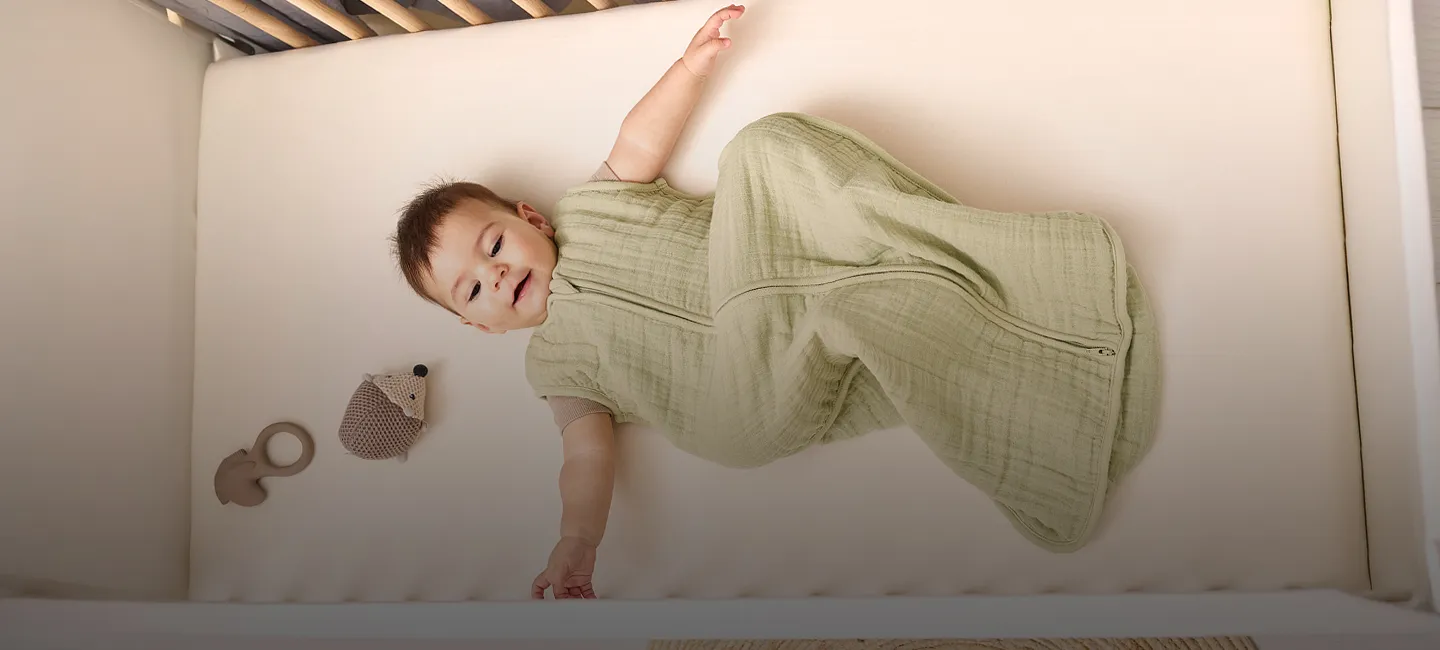 Baby im grünen Schlafsack liegt mit einem Teddybären und einem Beißring im Kinderbett und schaut nach oben.