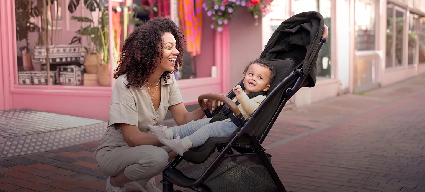 Eine Frau lächelt ein Baby in einem Kinderwagen auf einem Bürgersteig an; im Hintergrund sind rosafarbene Schaufenster und Pflanzen zu sehen.