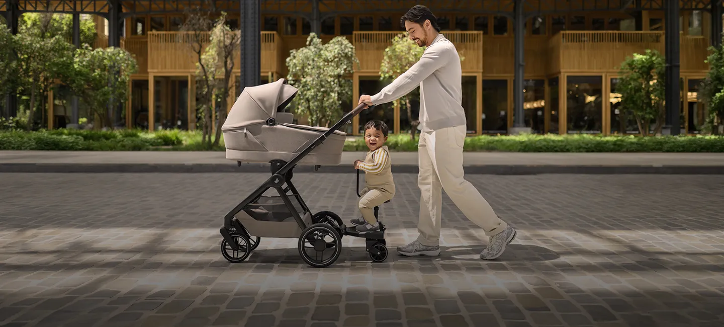 Ein Mann schiebt einen Kinderwagen, auf dem ein Kleinkind auf einem Mitfahrbrett steht; beide lächeln. Die Straße ist mit Kopfsteinpflaster belegt, im Hintergrund sind Grünanlagen und Gebäude zu sehen.