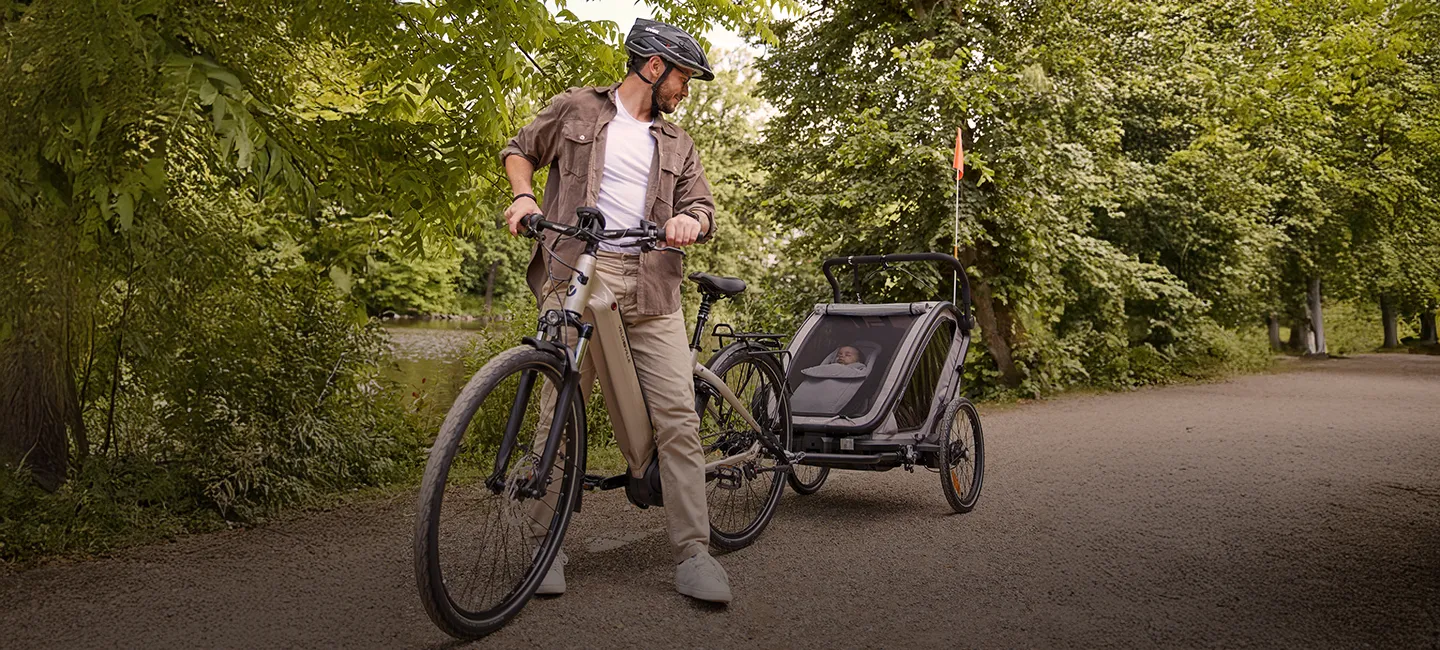 Ein Mann mit Fahrrad und Kinderanhänger steht auf einem von Bäumen gesäumten Weg und blickt zurück.
