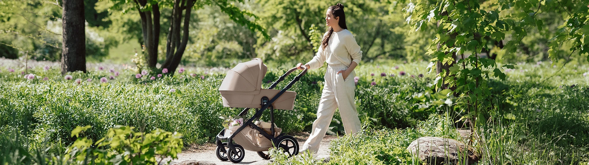 Eine Frau schlendert mit einem beigen Kinderwagen auf einem Pfad durch einen üppig grünen Park, umgeben von Bäumen und blühenden Blumen.