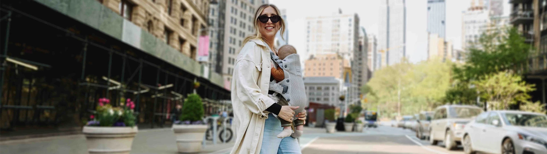 Eine Frau mit Sonnenbrille trägt ein Baby in einer Stadtlandschaft mit hohen Gebäuden, Grünflächen und Autos im Hintergrund an einem sonnigen Tag.
