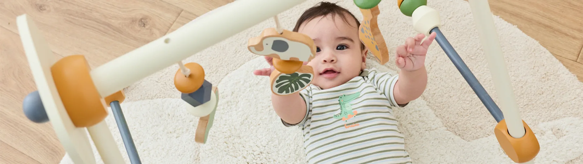 Ein Baby, das auf einem weißen Teppich liegt, greift nach einem hängenden Mobile mit Tierfiguren aus Holz und trägt ein gestreiftes Hemd mit Alligatormotiv.