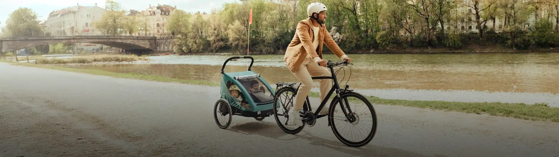 Eine Person in beigem Outfit und Helm fährt mit einem Kind im Anhänger auf einem Fahrrad einen Flussweg entlang, im Hintergrund sind eine Brücke und Bäume zu sehen.