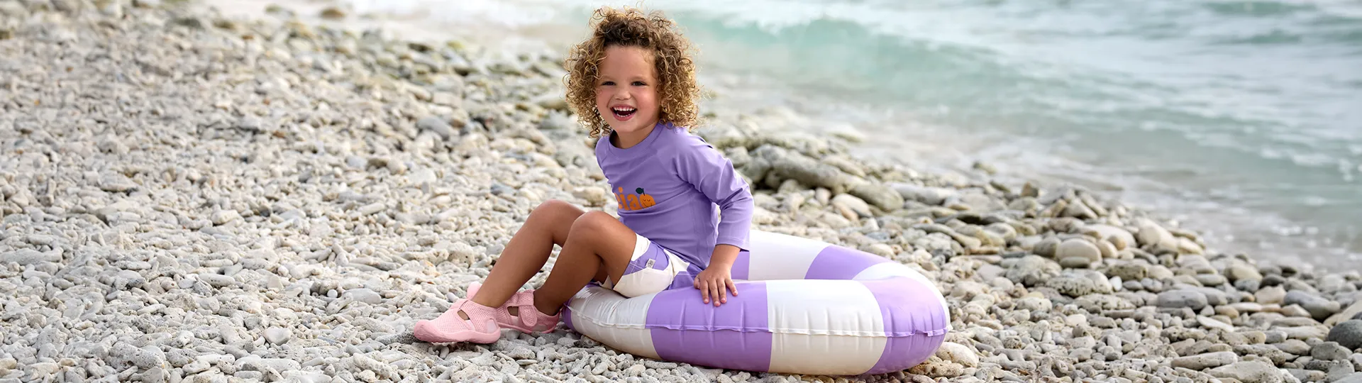 Ein Kind in lila Kleidung sitzt auf einer gestreiften Luftmatratze an einem felsigen Strand und lächelt mit dem Meer im Hintergrund.