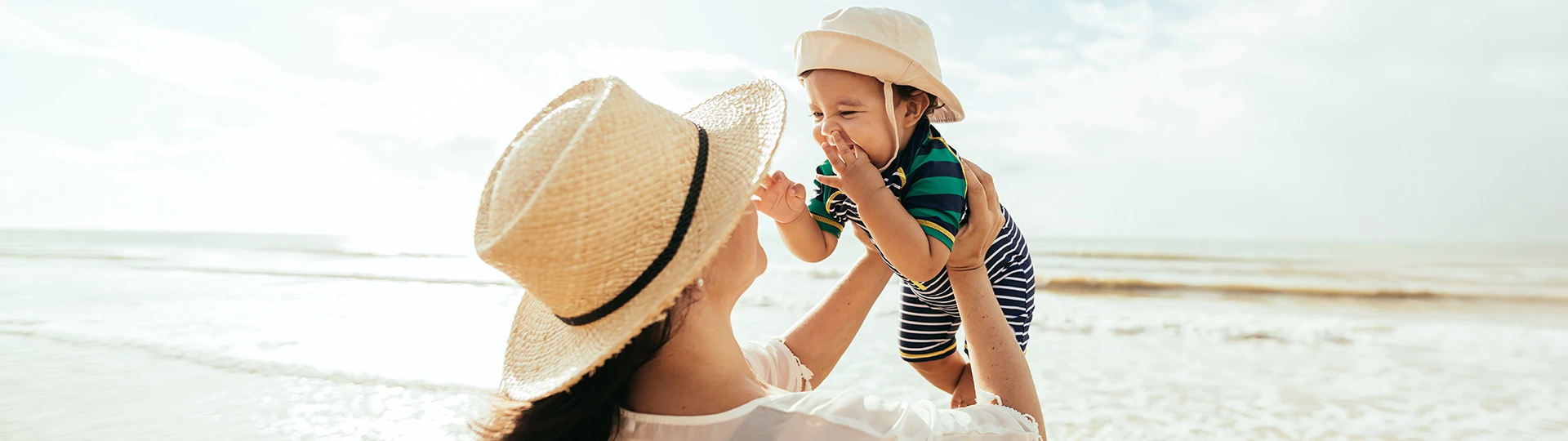 Eine Frau mit Sonnenhut hebt ein lächelndes Baby, das ebenfalls einen Hut und ein gestreiftes Outfit trägt, vor einer sonnigen Strandkulisse hoch.