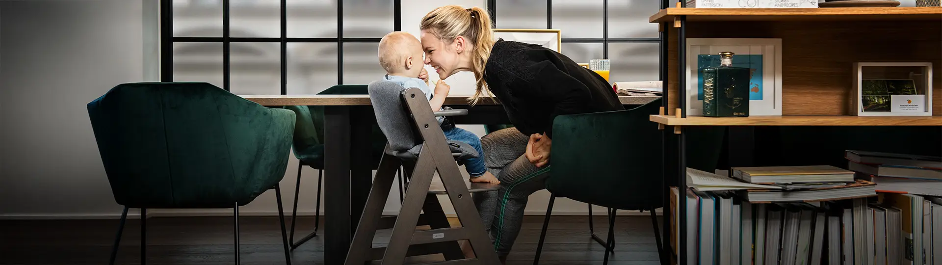 Eine Frau beugt sich über einen Esstisch und berührt spielerisch die Nase eines Babys, das in einem Hochstuhl sitzt, umgeben von Büchern und Regalen.