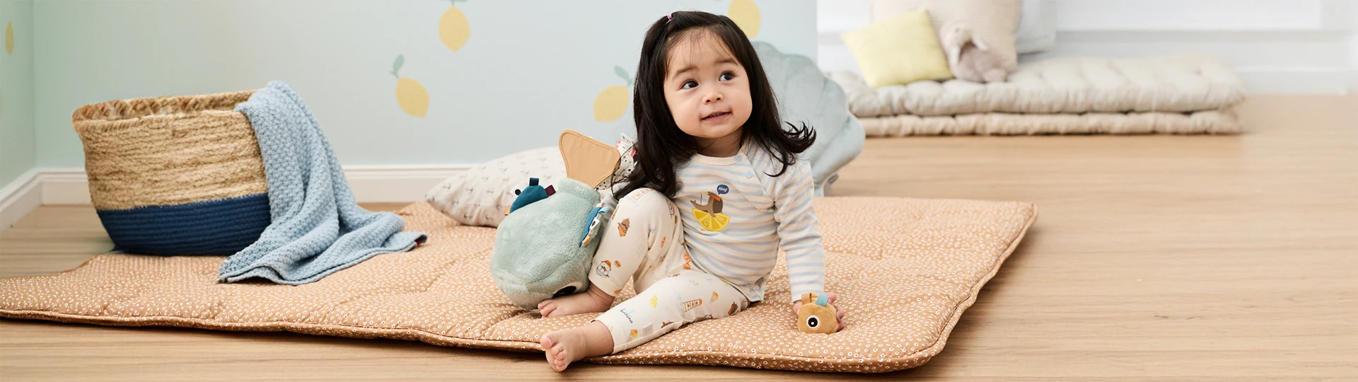 Ein Kleinkind sitzt auf einer weichen Matte in einem Kinderzimmer, umgeben von Stofftieren, Kissen und einem dekorativen Korb, im Hintergrund eine Wand mit Zitronenmuster.