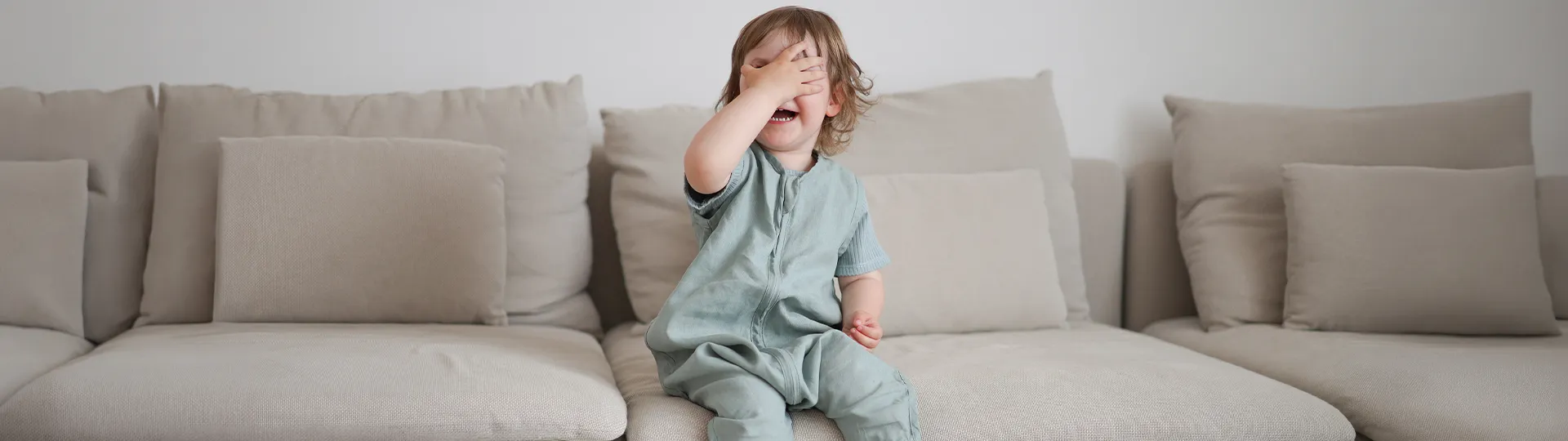 Ein Kind in hellblauer Kleidung sitzt lachend auf einem beigen Sofa und verdeckt mit einer Hand sein Gesicht.