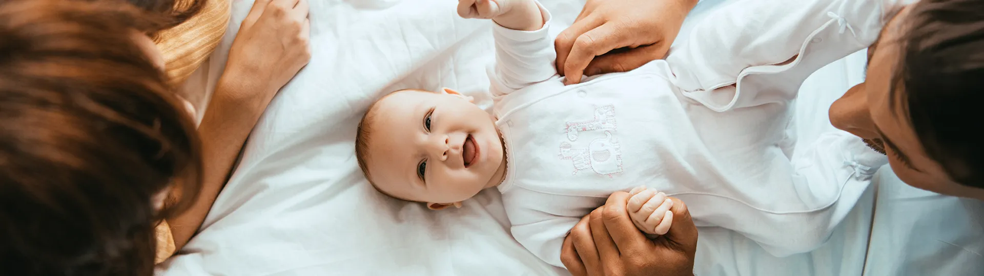 Ein lächelndes Baby liegt auf einem Bett, umgeben von Erwachsenen, und trägt einen weißen Strampler mit einer aufgestickten Giraffe.