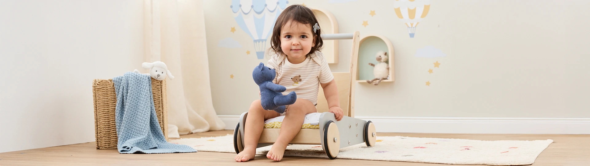 Ein Kleinkind sitzt auf einem hölzernen Schaukelstuhl und hält einen blauen Teddybären in einem Kinderzimmer mit Heißluftballon-Wanddekoration und einem geflochtenen Korb.