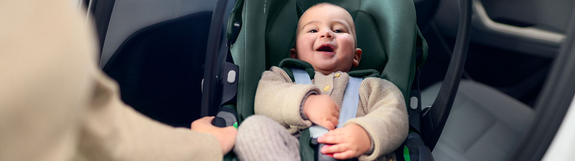 Ein glückliches Baby sitzt lächelnd in einem Kindersitz. Eine Erwachsenenhand justiert den Sicherheitsgurt. Im Hintergrund ist der Innenraum des Autos zu sehen.