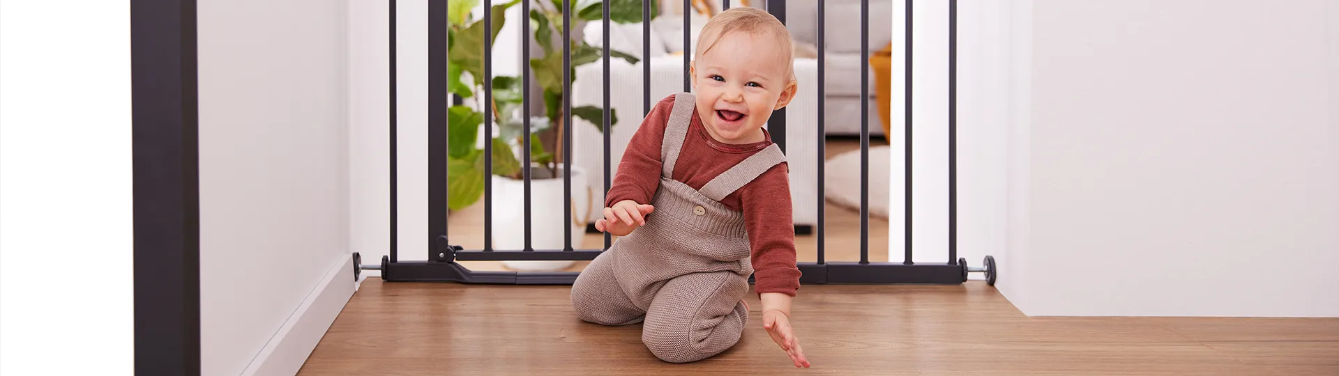 Ein lächelndes Baby in einem roten Hemd und beigen Overalls krabbelt auf einem Holzboden in der Nähe eines Sicherheitsgitters; im Hintergrund sind Pflanzen und Möbel zu sehen.