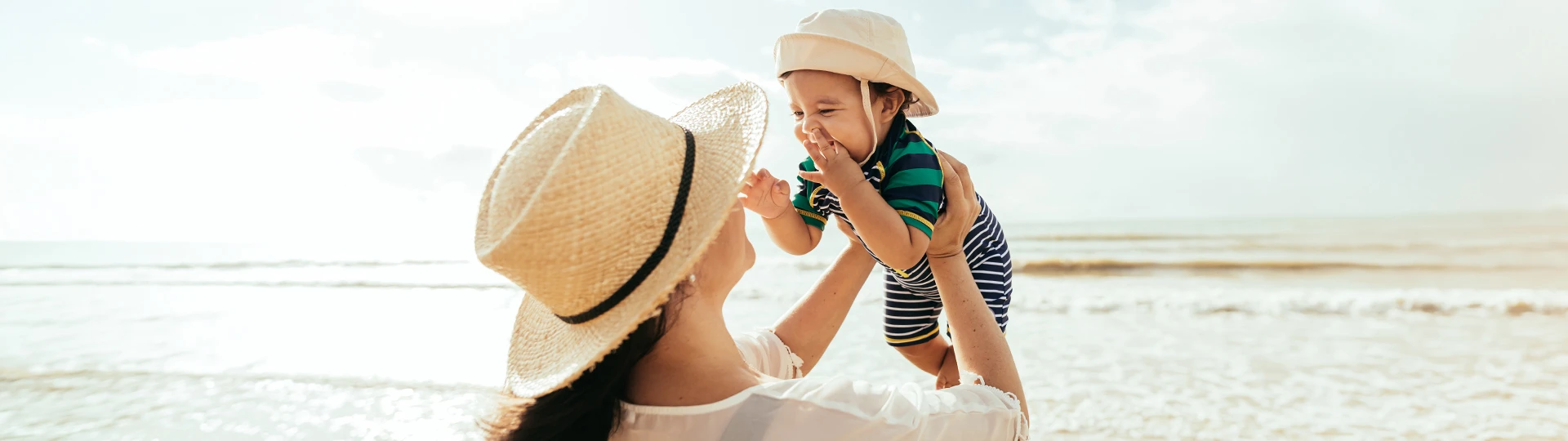 Eine Frau mit Strohhut hebt am Strand ein lächelndes Baby hoch, das einen Sonnenhut und ein gestreiftes Outfit trägt; im Hintergrund ist das Meer zu sehen.