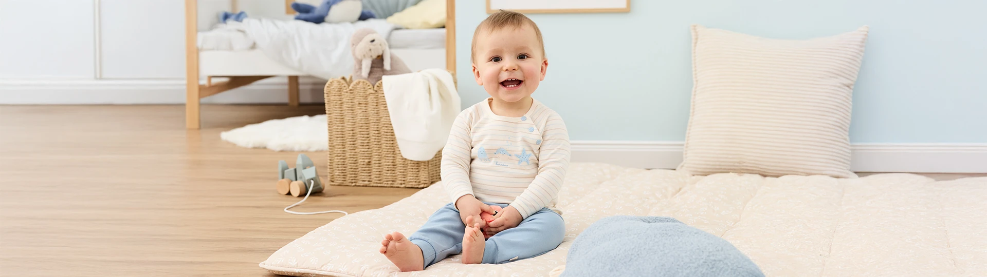 Ein lächelndes Baby sitzt auf einer weichen Spielmatte in einem gemütlichen Zimmer, in der Nähe stehen ein Weidenkorb und Spielzeug.