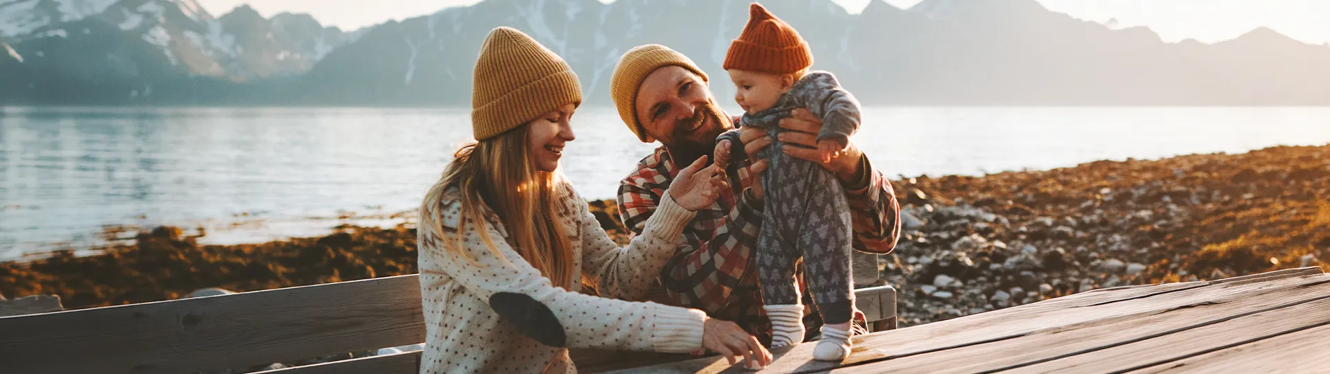 Eine Familie sitzt in warmer Kleidung auf einer Holzbank am See – im Hintergrund schneebedeckte Berge – und lächelt ihr Baby an.